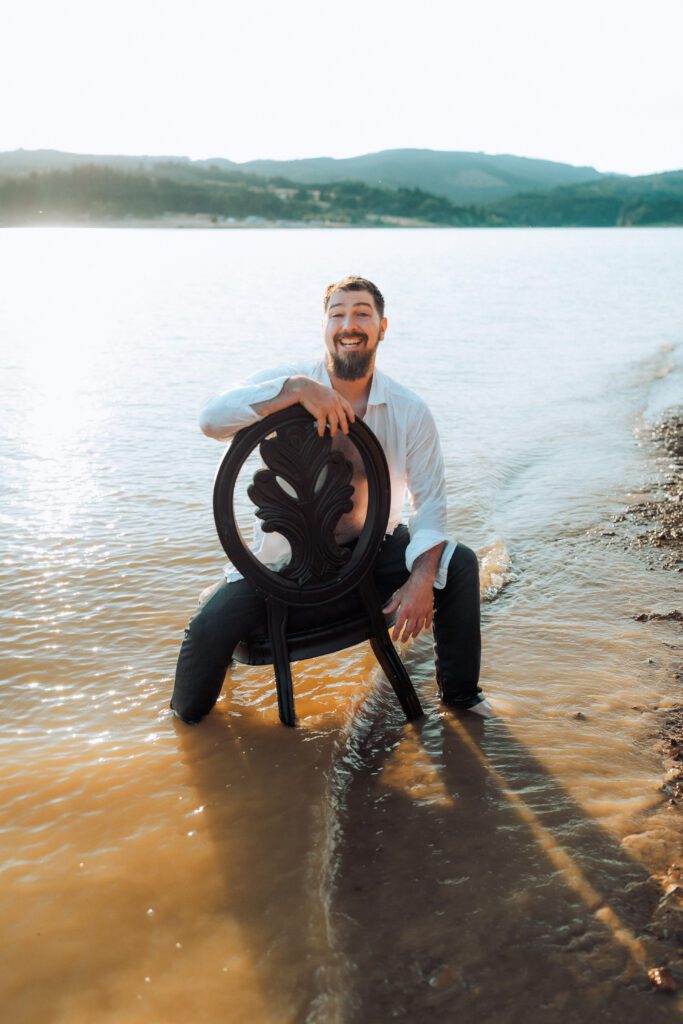 Man sitting on a dark chair in shallow lake water, smiling during a relaxed brand session in the Pacific Northwest