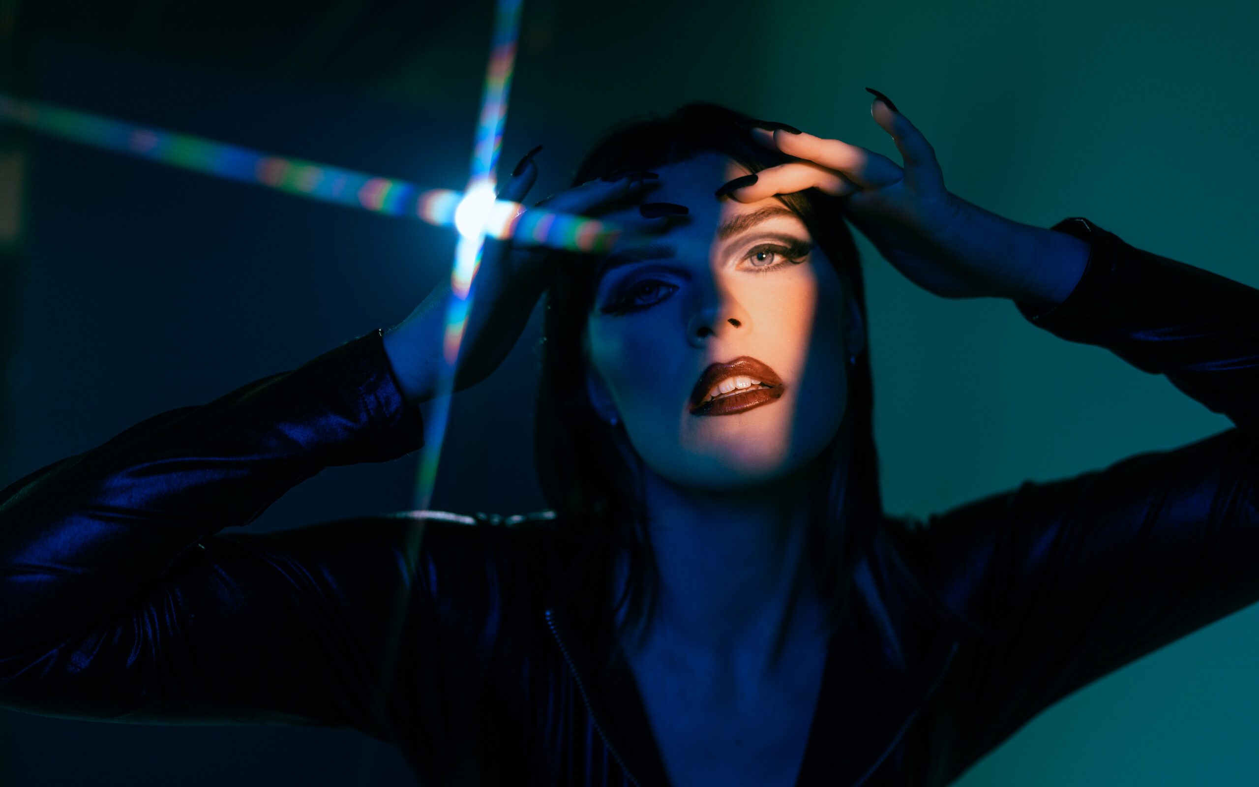 Dramatic studio portrait of a woman in dark clothing with hands framing her face, lit by bold blue-green lighting and a prism flare—captured during a creative session inside The Studio Lab by Mewphotovideo