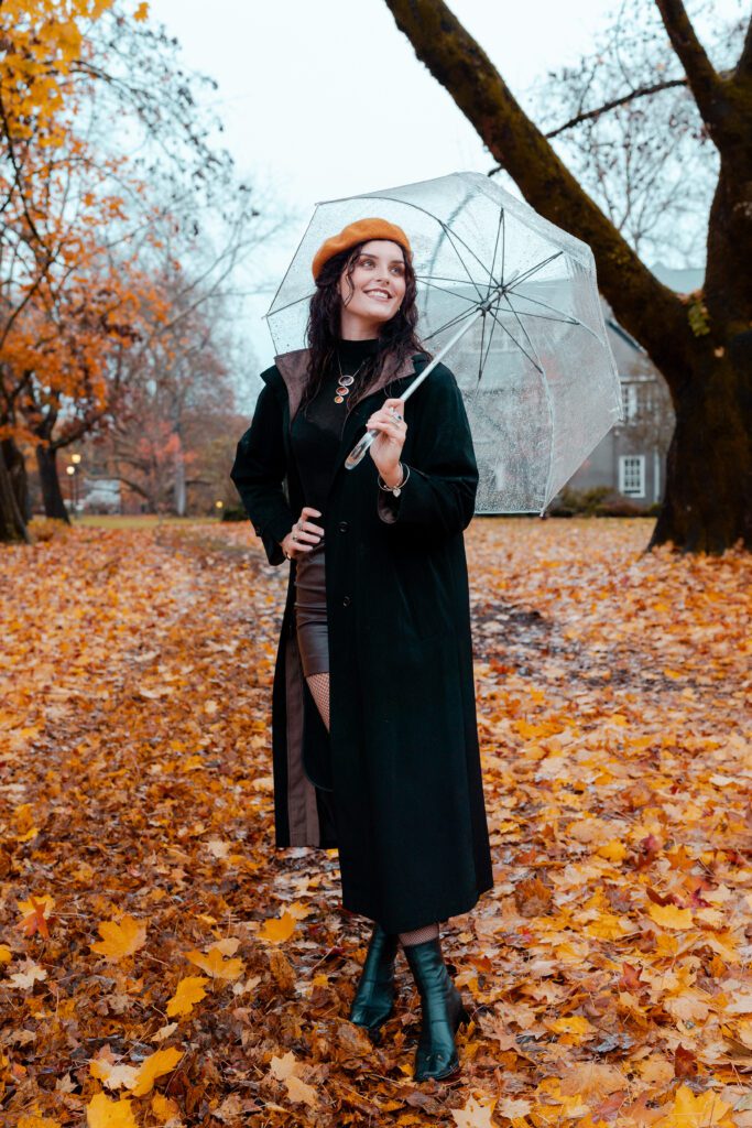 Woman in black trench coat standing confidently among orange leaves and bare trees, blending elegance and edge in an Outdoor Adventure session during fall in Portland