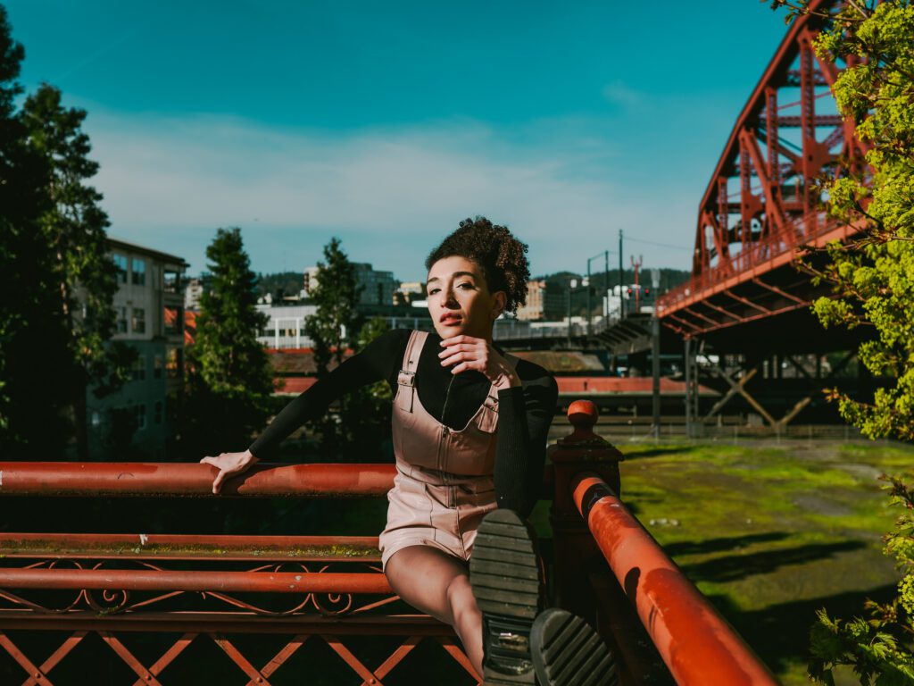 Creative brand portrait of a woman seated on a red bridge railing in Portland, Oregon, with industrial architecture in the background and confident body language