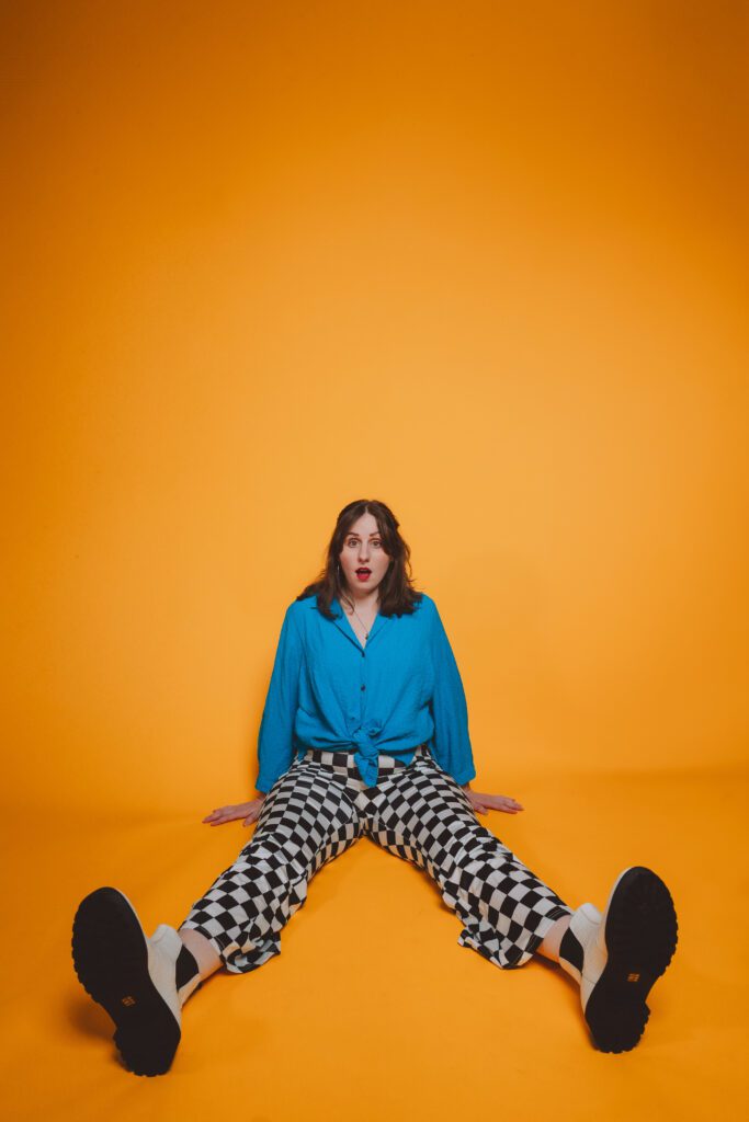 Portland portrait of a woman sitting on an orange backdrop wearing a bright blue top and checkered pants, captured in a modern, colorful studio setup.