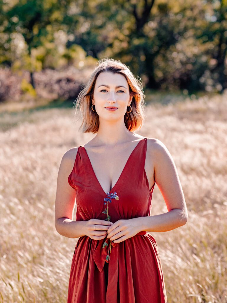 Elegant woman in a red dress holding wildflowers, standing in tall grass under warm light—captured during a nature-based Outdoor Adventure session in central Texas