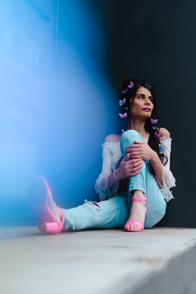 Creative personal brand photo of a woman sitting thoughtfully against a textured wall, surrounded by soft blue light, butterflies, and pastel styling.