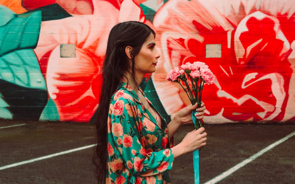 Side profile branding photo of a woman holding pink carnations in front of a bold red and teal mural in Portland, wearing a matching floral outfit—perfect for creative professionals seeking vibrant personal brand imagery.