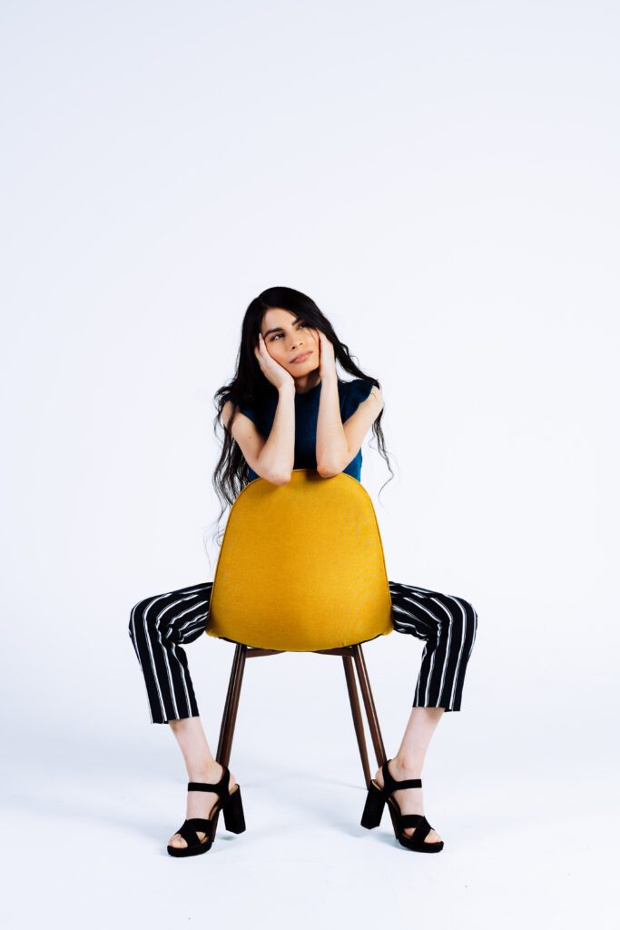 Editorial portrait with contrast styling, featuring a bright yellow chair and strong seated pose.