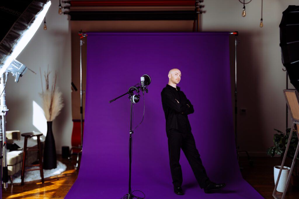 Man in black clothing standing confidently in front of a purple backdrop with a microphone setup, styled for a brand video and photo session in Speak Your Language by Mewphotovideo