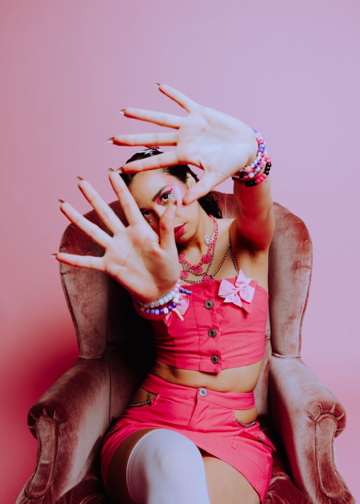 Model in a hot pink outfit raises her hands to frame the camera lens during a bold monochrome studio portrait session in PDX, styled with beaded jewelry and expressive pink makeup.