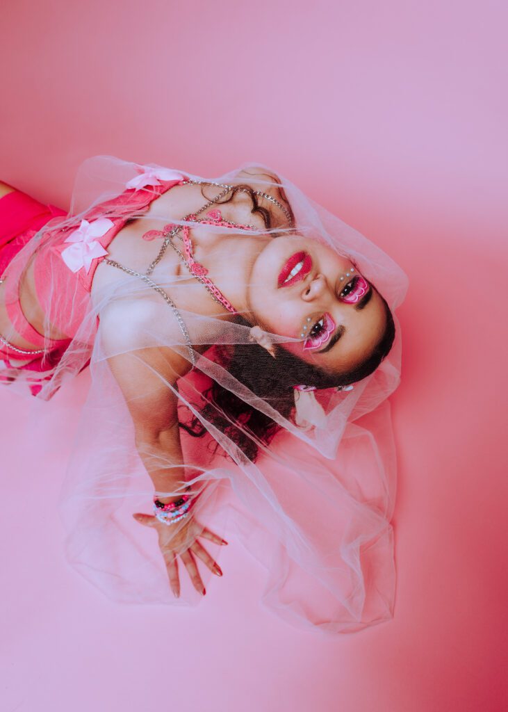 Creative portrait of model lying on a pink studio floor wearing a sheer veil and dramatic eye makeup, captured during a monochrome bubblegum shoot at Cobalt Studios PDX.