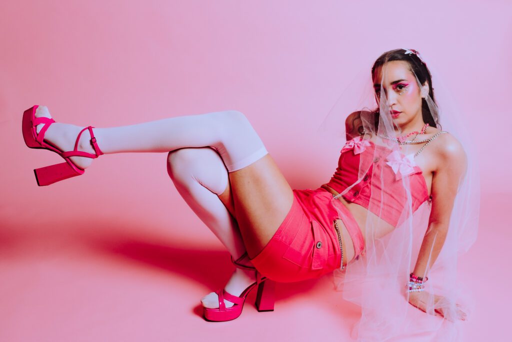 Monochrome portrait of model wearing a pink two-piece outfit and sheer veil, seated against a pink backdrop with bold styling and accessories in a creative studio session.