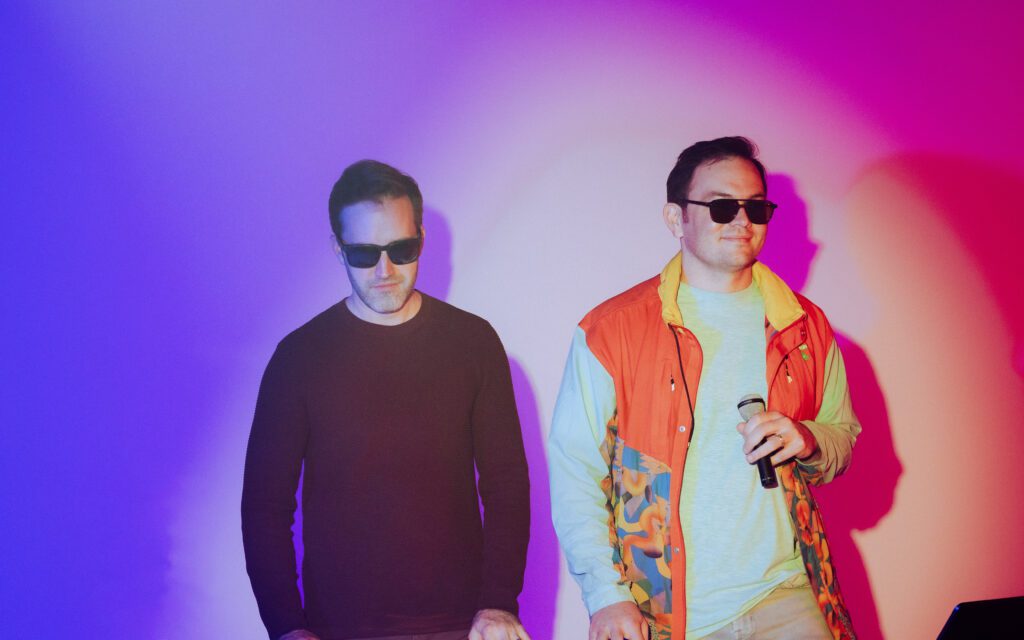Househusband members wearing sunglasses and holding a mic under pink and purple lighting at Mewphotovideo's Portland studio