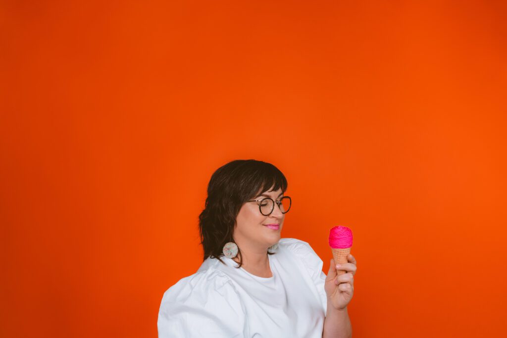 Woman in white top smiling while holding a pink scoop of ice cream against a bold orange background, captured during a playful and personality-filled Speak Your Language session by Mewphotovideo
