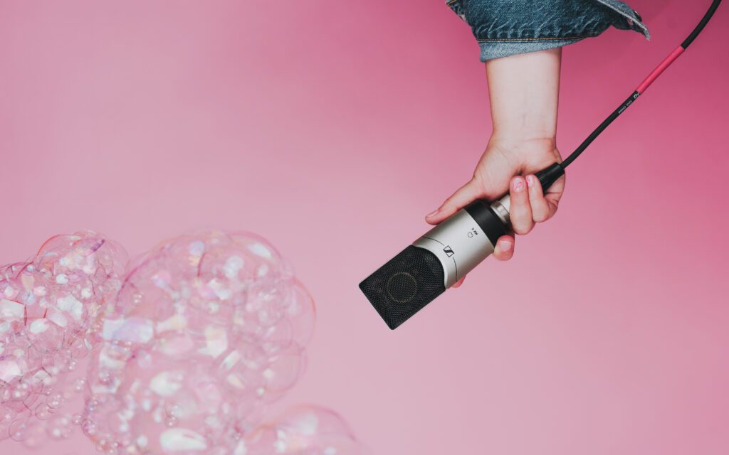Close-up of hand holding a studio microphone with iridescent bubbles rising against a soft pink background—playful and symbolic imagery for brand video storytelling