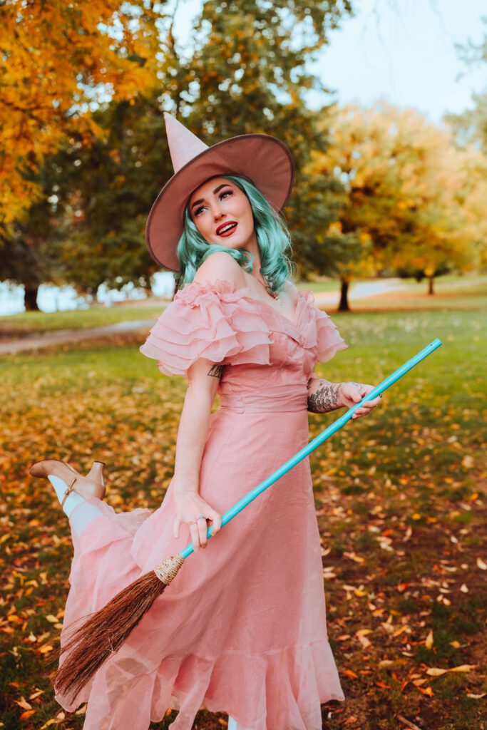 Creative portrait of a woman in a pink witch dress and hat holding a broom in a Portland park during fall, captured by a creative portrait photographer in Portland.