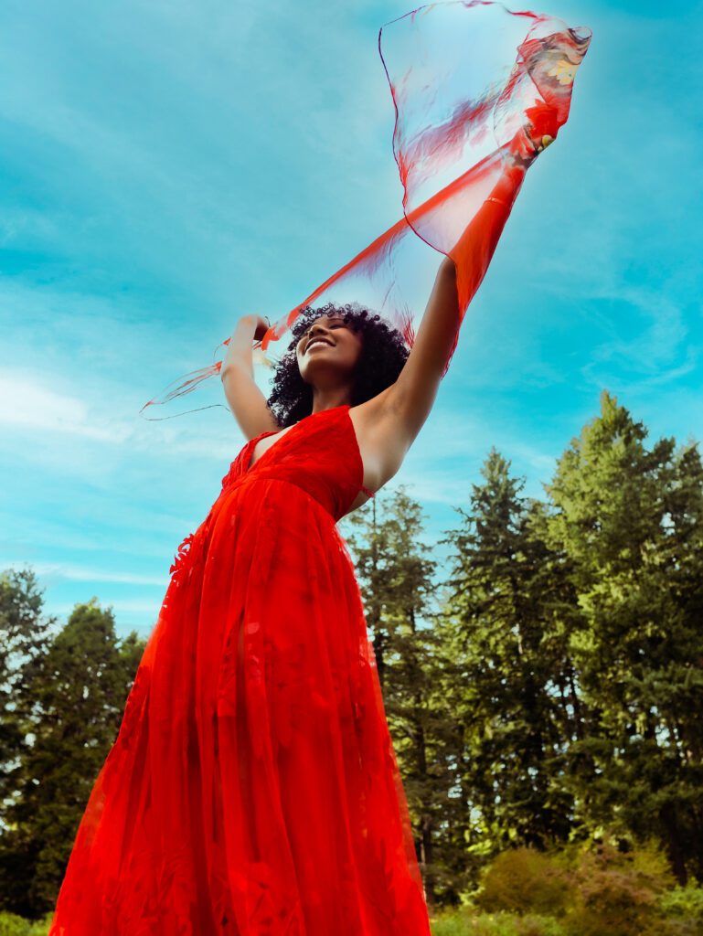 Client in a flowing red dress holding fabric overhead under bright blue sky and surrounded by evergreens—capturing energy and freedom in an Outdoor Adventure brand session