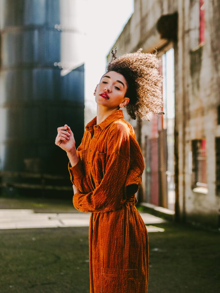 Woman in a rust-orange dress posing confidently in an industrial alleyway with dramatic backlight—part of an Outdoor Adventure brand shoot in Portland’s creative district