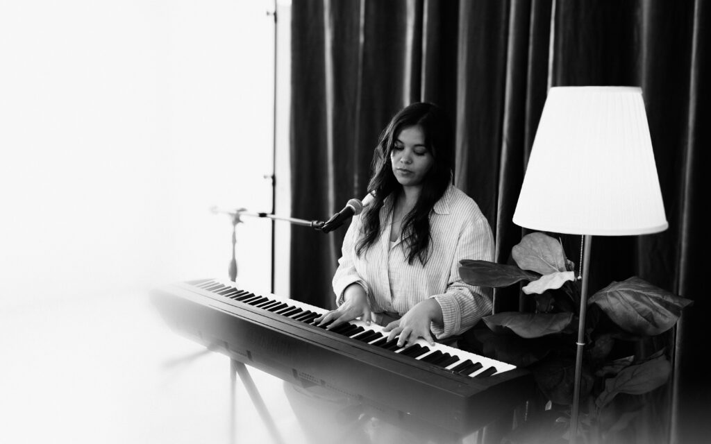 Black and white portrait of an artist playing keyboard beside a lamp in a creative studio setting