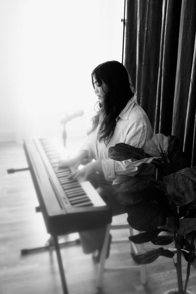Black and white portrait of a musician playing keyboard and singing into a microphone in soft natural light