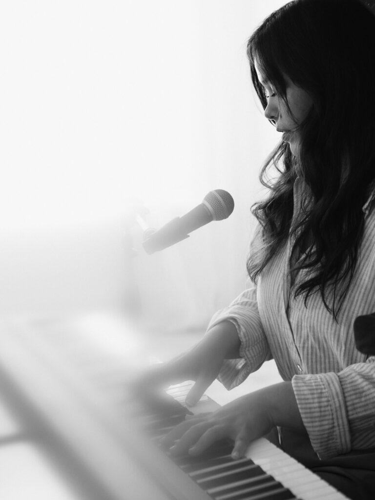 Black and white portrait of a musician playing keyboard and singing into a microphone in soft natural light