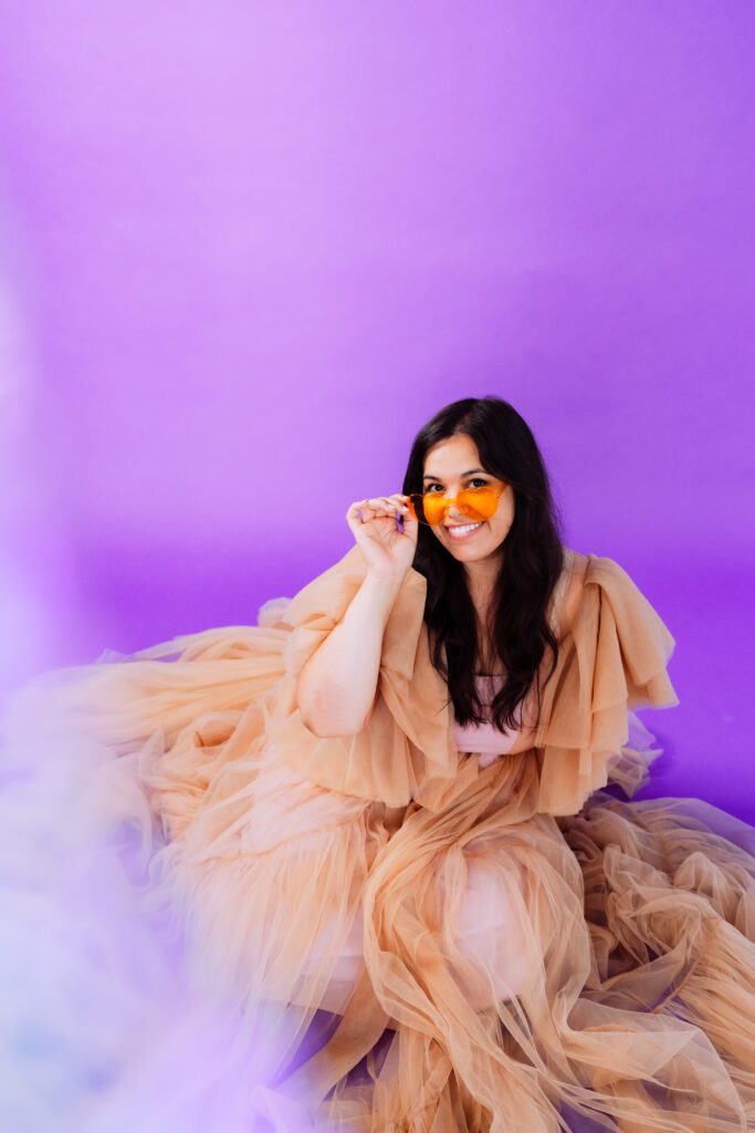 Artist playfully posing in layered peach gown and sunglasses during a monochrome brand shoot in studio