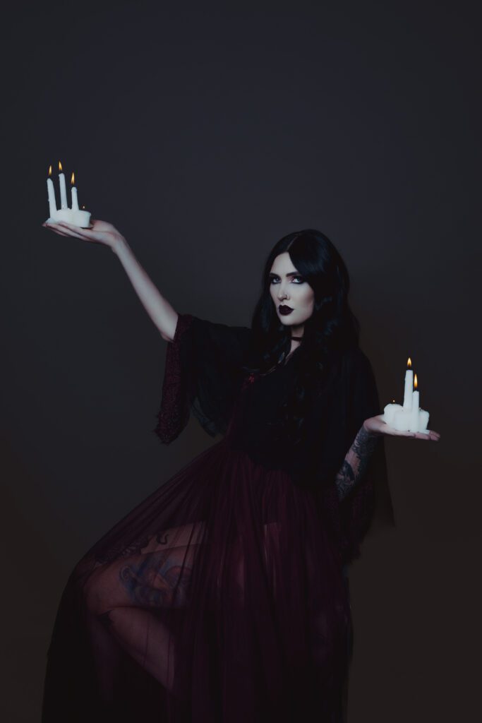 Gothic-inspired studio portrait of a woman in sheer burgundy gown holding lit white candles in both hands against a dark backdrop—captured during a moody, cinematic session inside The Studio Lab by Mewphotovideo