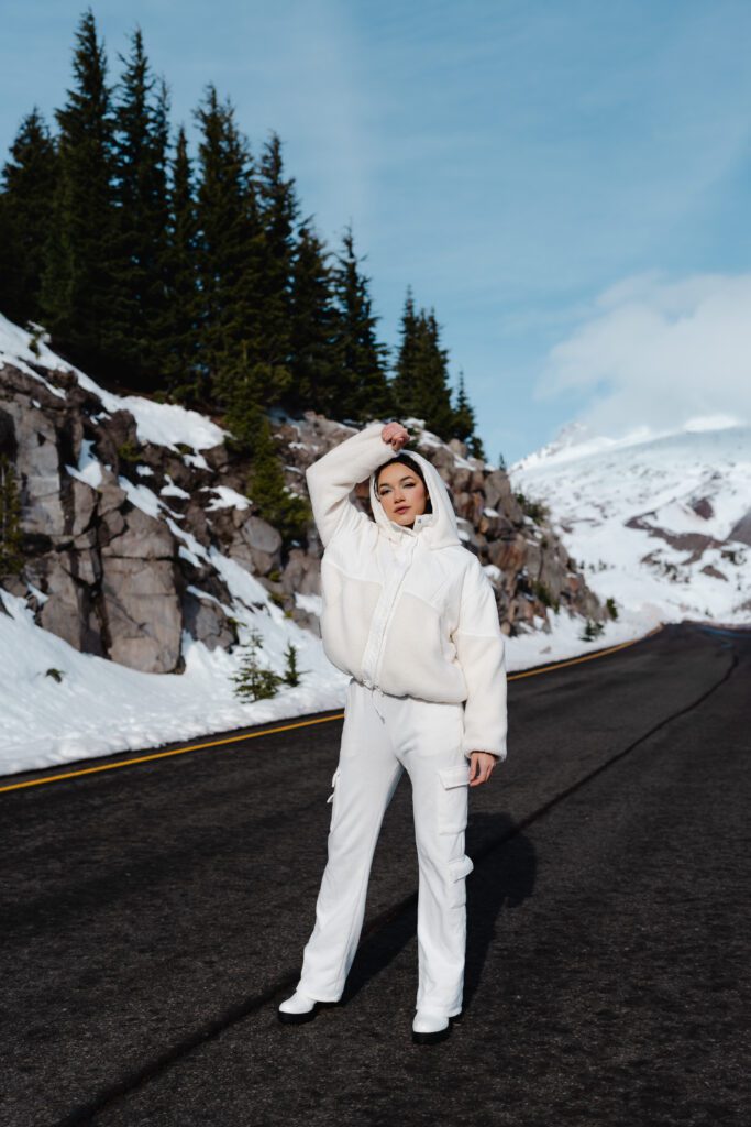 Client in an all-white outfit standing confidently on a snow-lined mountain road, framed by pine trees and sky—part of a bold winter Outdoor Adventure brand session in Oregon