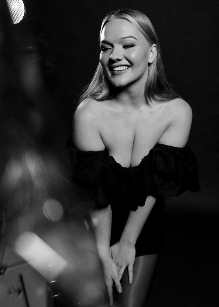 Candid black-and-white studio portrait of a woman in an off-shoulder dress laughing and leaning forward under soft spotlight, captured during a creative portrait session