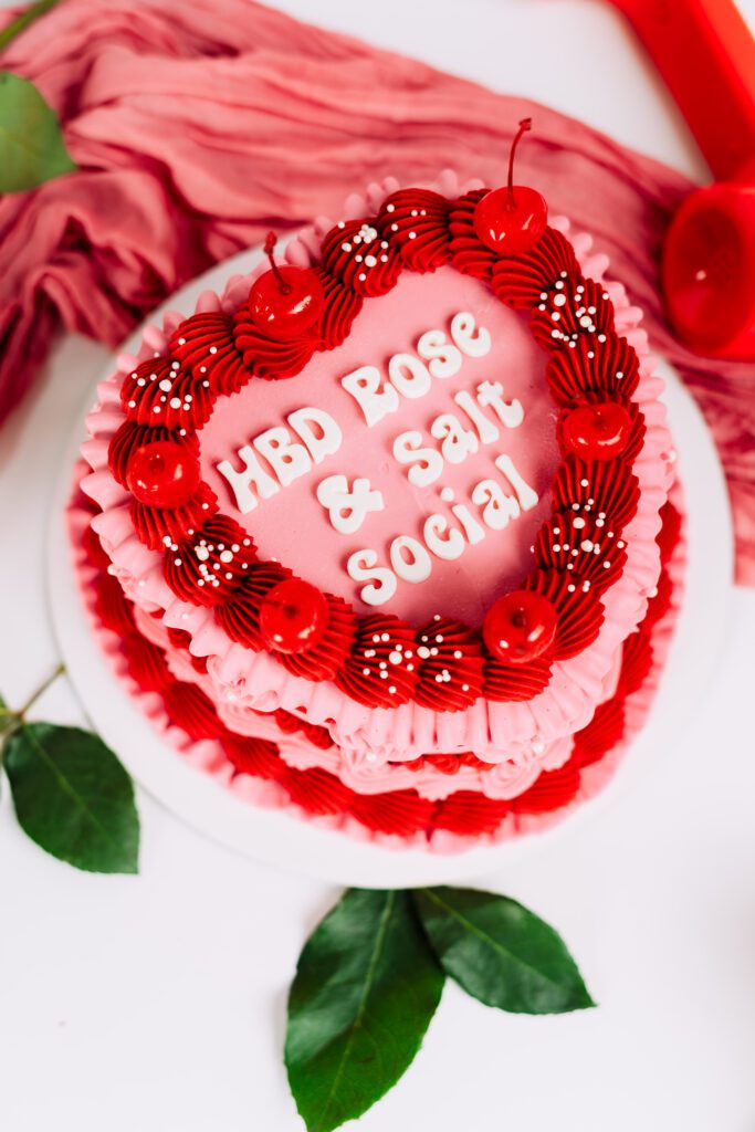 Overhead close-up of a custom pink and red heart-shaped cake with cherries and “HBD Rose & Salt Social” piped on top, styled for a brand's anniversary shoot