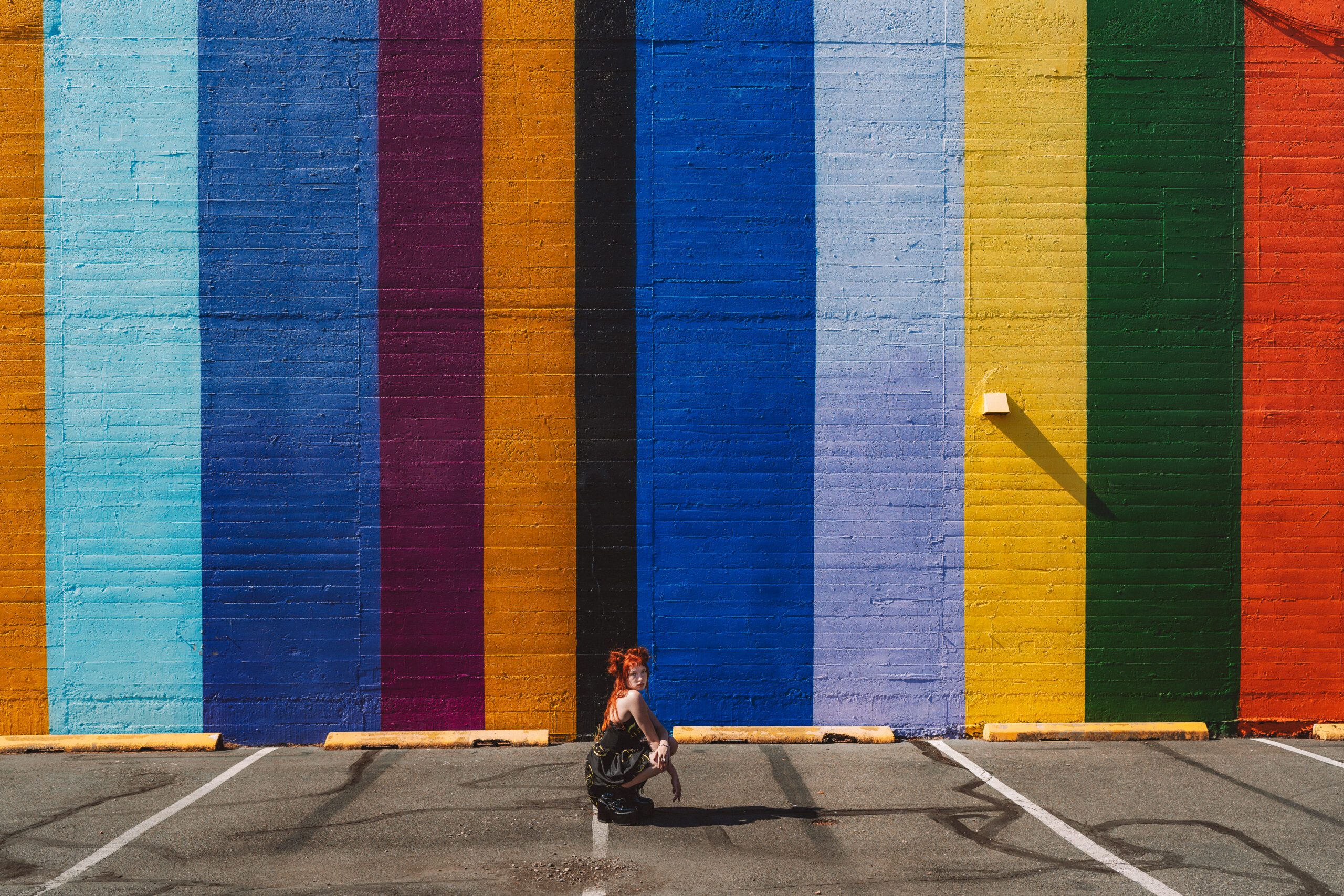 Creative portrait of a red-haired woman crouching in front of a vibrant rainbow-striped mural in a Portland parking lot, captured by a Portland creative portrait photographer.
