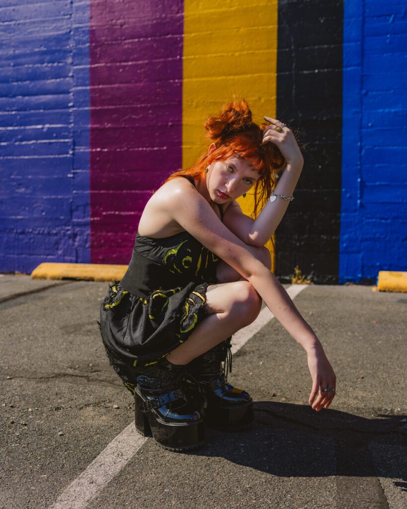 Woman crouching confidently in front of a rainbow-painted brick wall, combining colorful background and expressive styling.