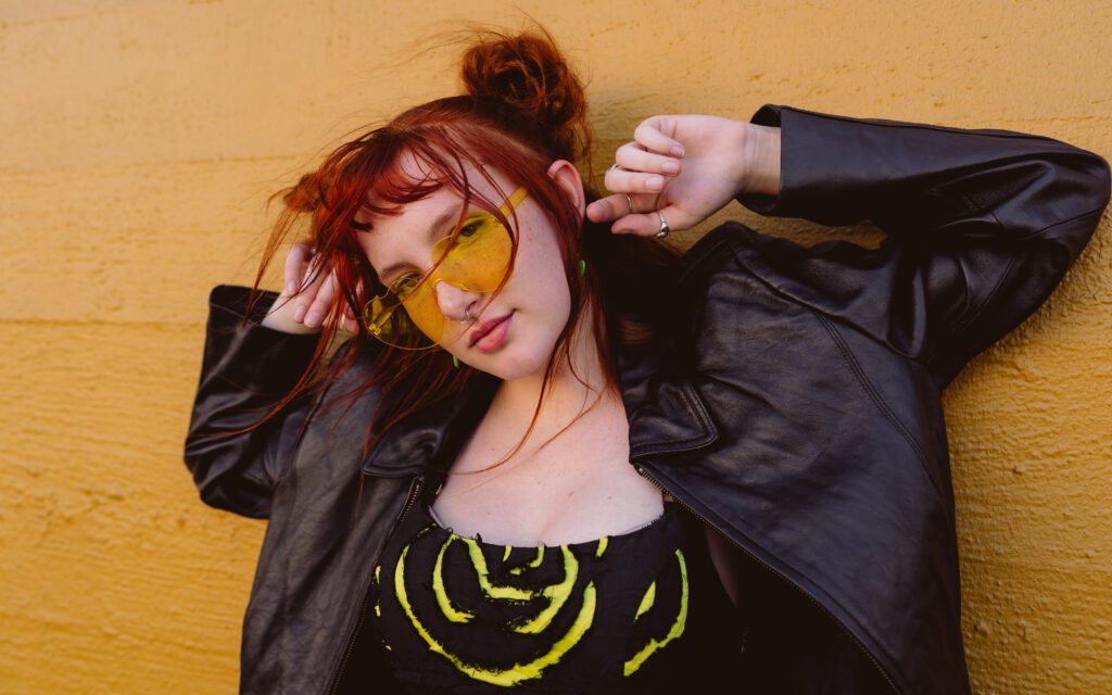 Creative close-up portrait of a woman in yellow sunglasses and leather jacket against a textured yellow brick wall during an Outdoor Adventure session.