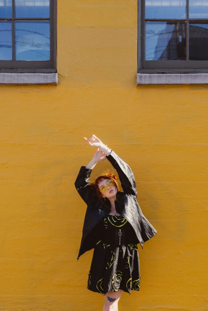 Artistic editorial shot of a woman posing with arms raised in front of a yellow wall with blue windows, showcasing movement and energy in branding photography by Mewphotovideo