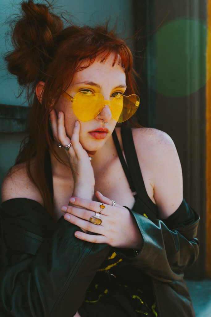 Woman resting her face in her hand, wearing yellow sunglasses and a black jacket in front of a teal wall during a creative portrait session.