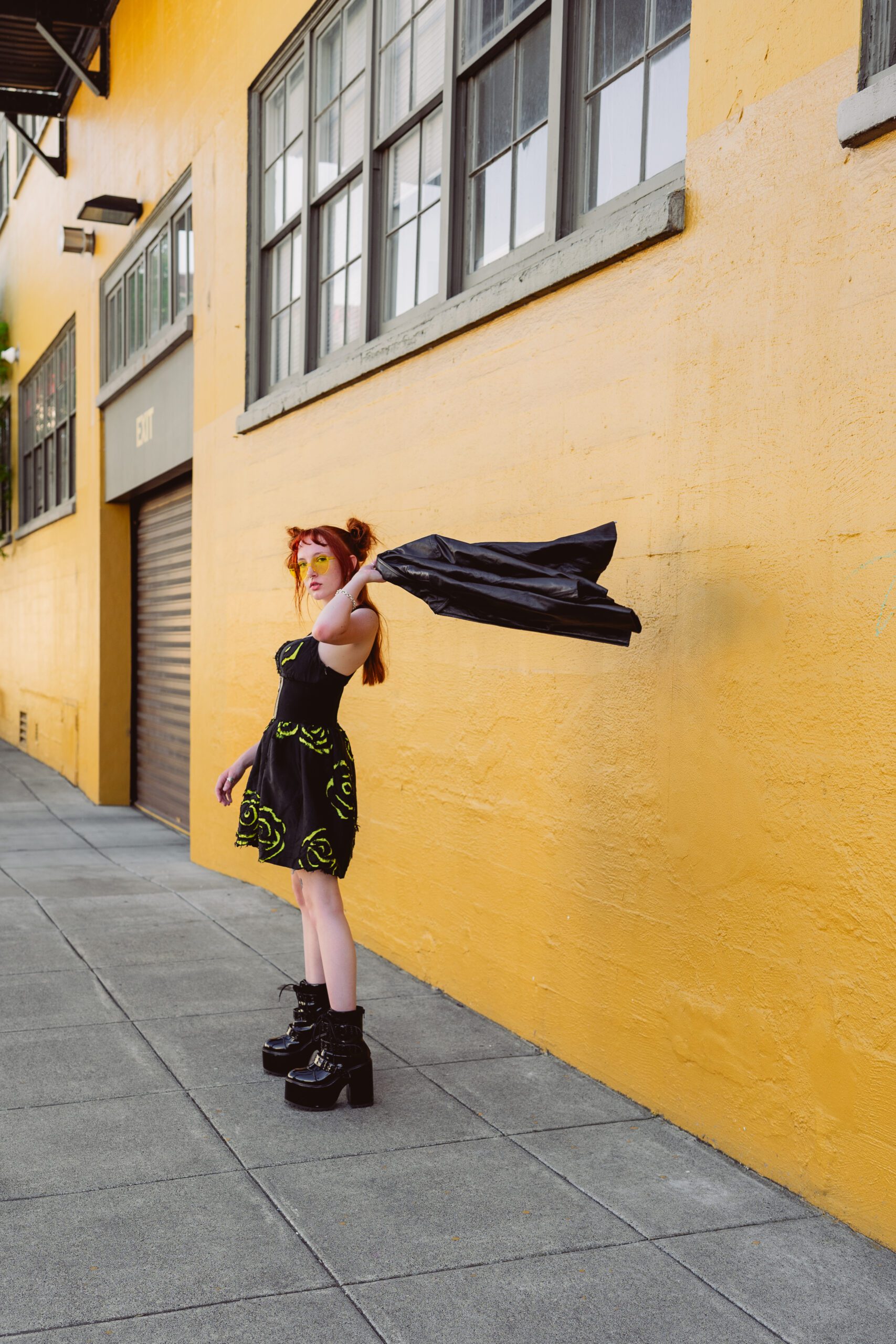 Woman throwing a black jacket mid-air while posing confidently against a mustard yellow wall during a dynamic summer portrait shoot.