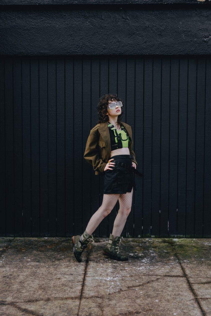 Portrait of a model in cyberpunk streetwear standing confidently in front of a black wooden wall in Portland, taken by a bold creative portrait photographer.