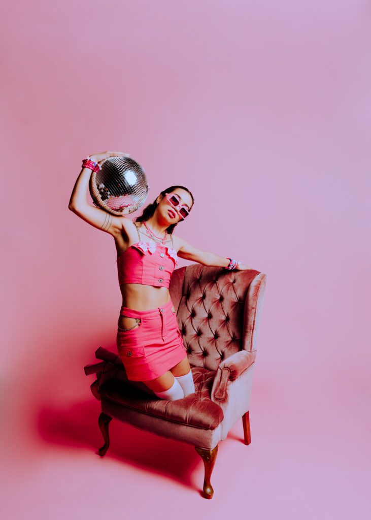 Creative portrait of model kneeling on a vintage velvet chair in a pink studio set, wearing bold platform heels and a hot pink outfit, captured during a pink monochrome photoshoot at Cobalt Studios.